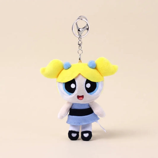 A plush keychain charm of the Powerpuff Girls character Bubbles, featuring yellow hair, a blue and black striped dress, and a silver metal key ring, set against a solid beige background.