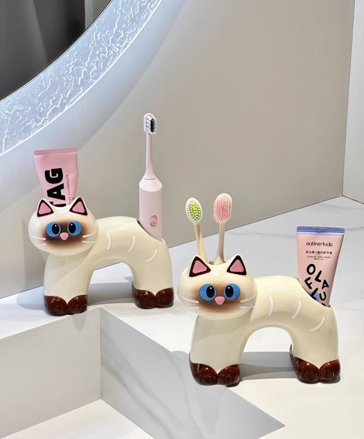 Two ceramic cat toothbrush and toothpaste holders in black and cream, sitting on a neutral tabletop.
