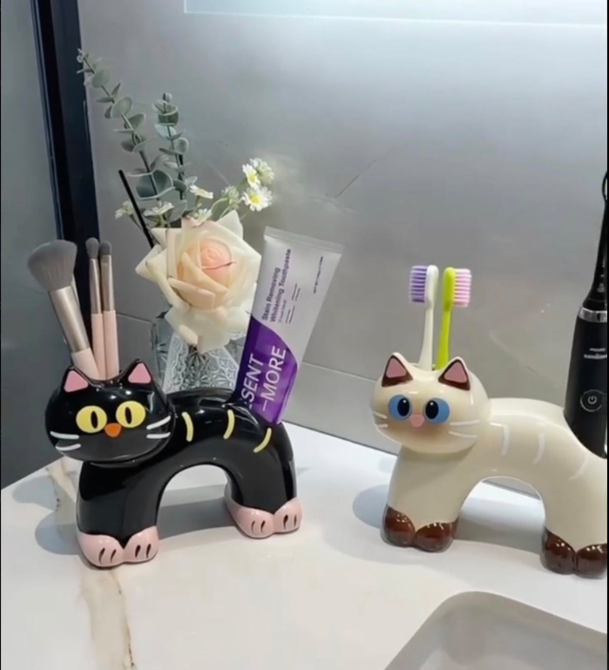 Two ceramic cat toothbrush and toothpaste holders in black and cream, sitting on a neutral tabletop.