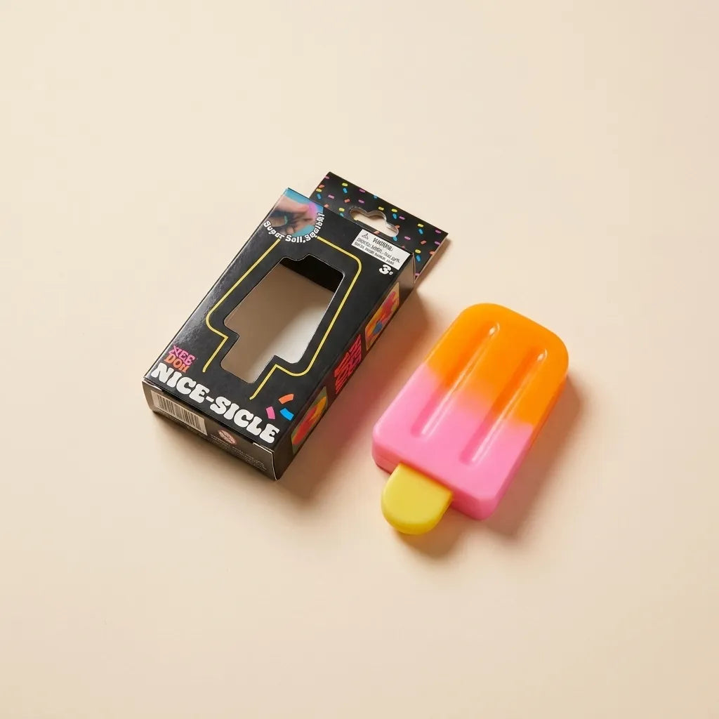 A novelty popsicle-shaped object with a yellow stick, pink-to-orange gradient body, shown next to its black retail box labeled 'NICE-SICLE' on a warm beige background.