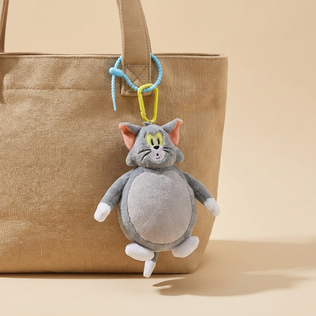 A plush character keychain shaped like a gray cat with pink ears, yellow eyes, and white paws, hanging from a blue and yellow clip on a beige canvas tote bag against a warm beige background.