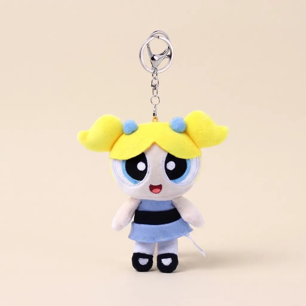 A plush keychain charm of the Powerpuff Girls character Bubbles, featuring yellow hair, a blue and black striped dress, and a silver metal key ring, set against a solid beige background.