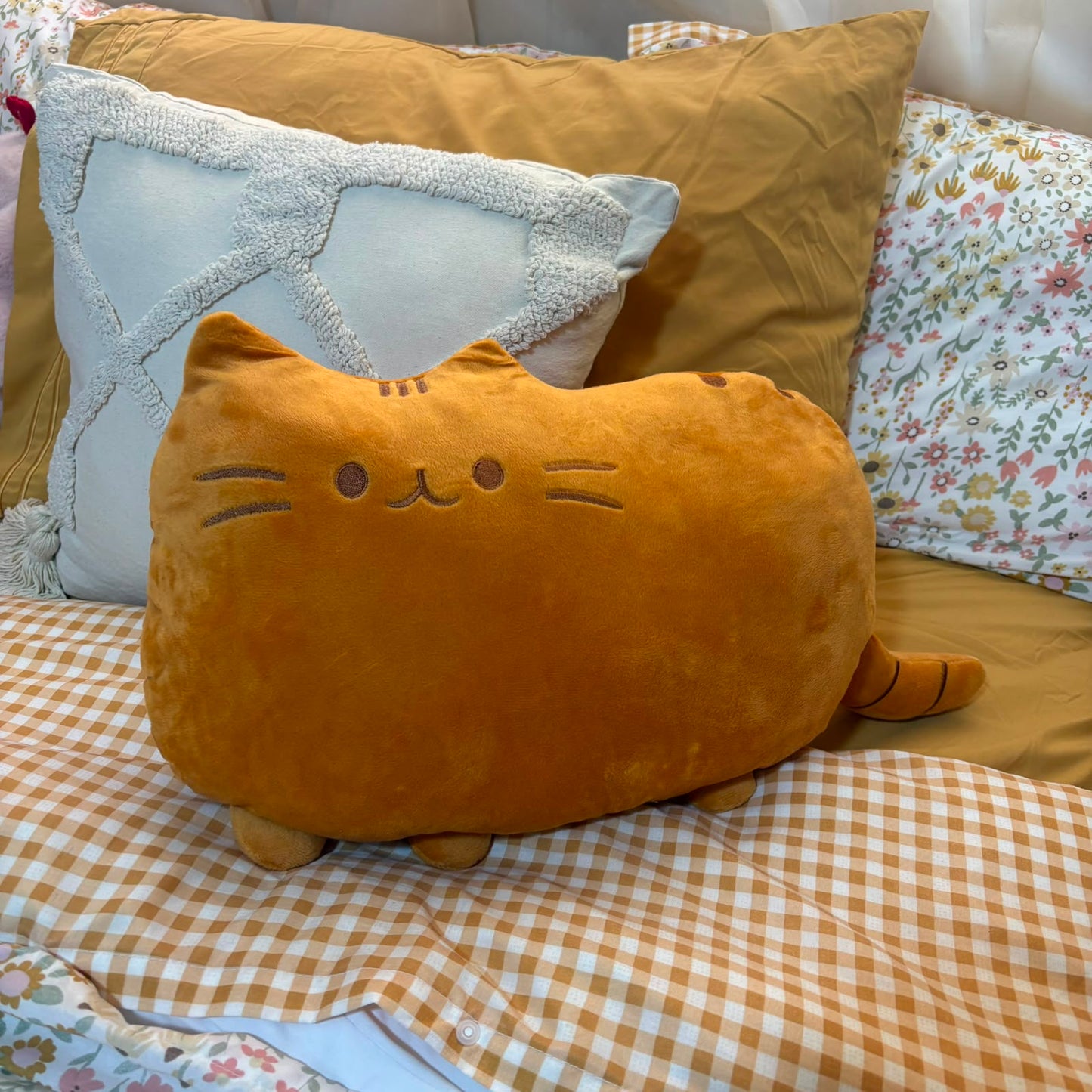 A plush cat-shaped pillow in a warm mustard-yellow color, styled on a bed with a brown and white gingham check blanket, a tan pillow, and a white patterned pillow in the background.