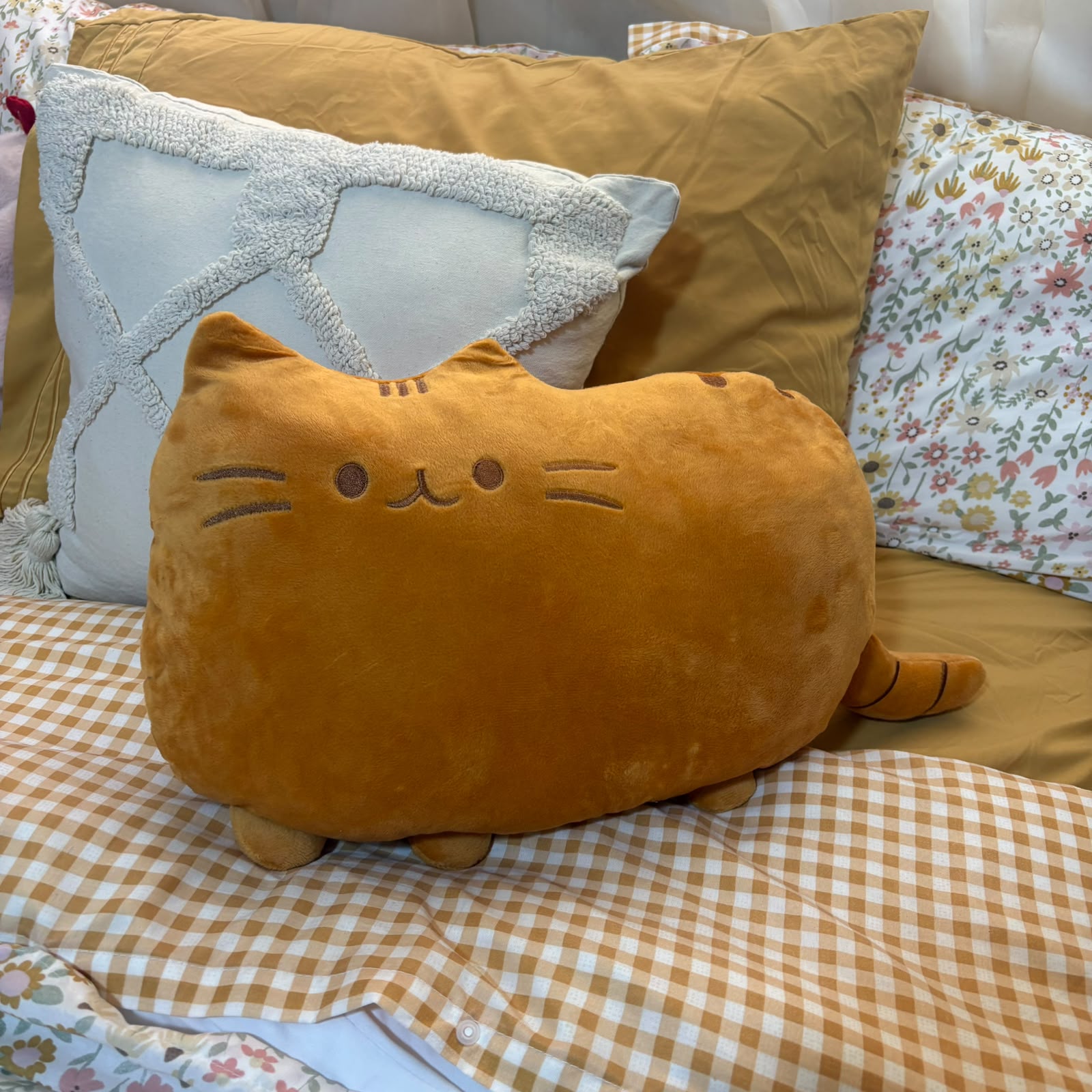 A plush cat-shaped pillow in a warm mustard-yellow color, styled on a bed with a brown and white gingham check blanket, a tan pillow, and a white patterned pillow in the background.