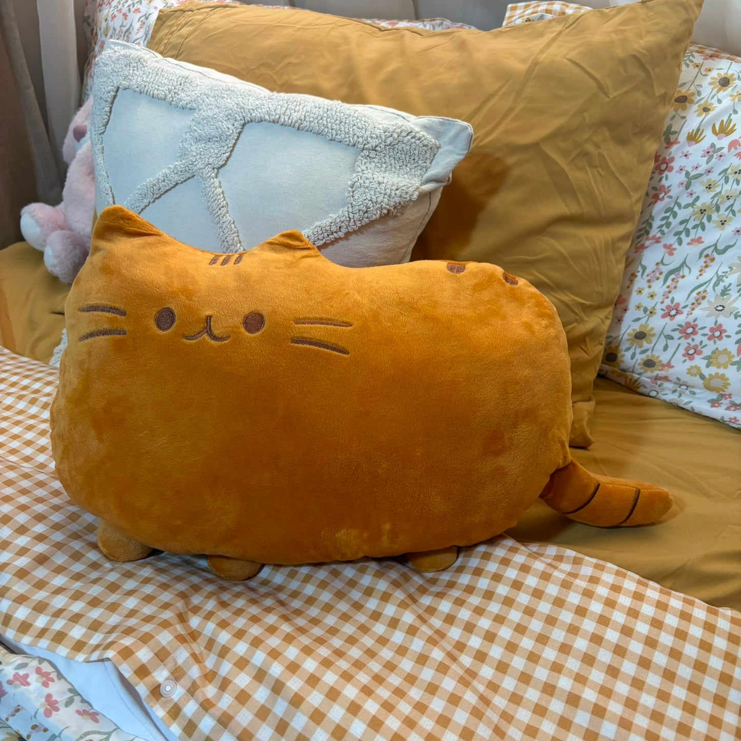 A plush cat-shaped pillow in a warm mustard-yellow color sits on a bed with a brown-and-white gingham blanket, a large tan pillow, and a light blue textured accent pillow.