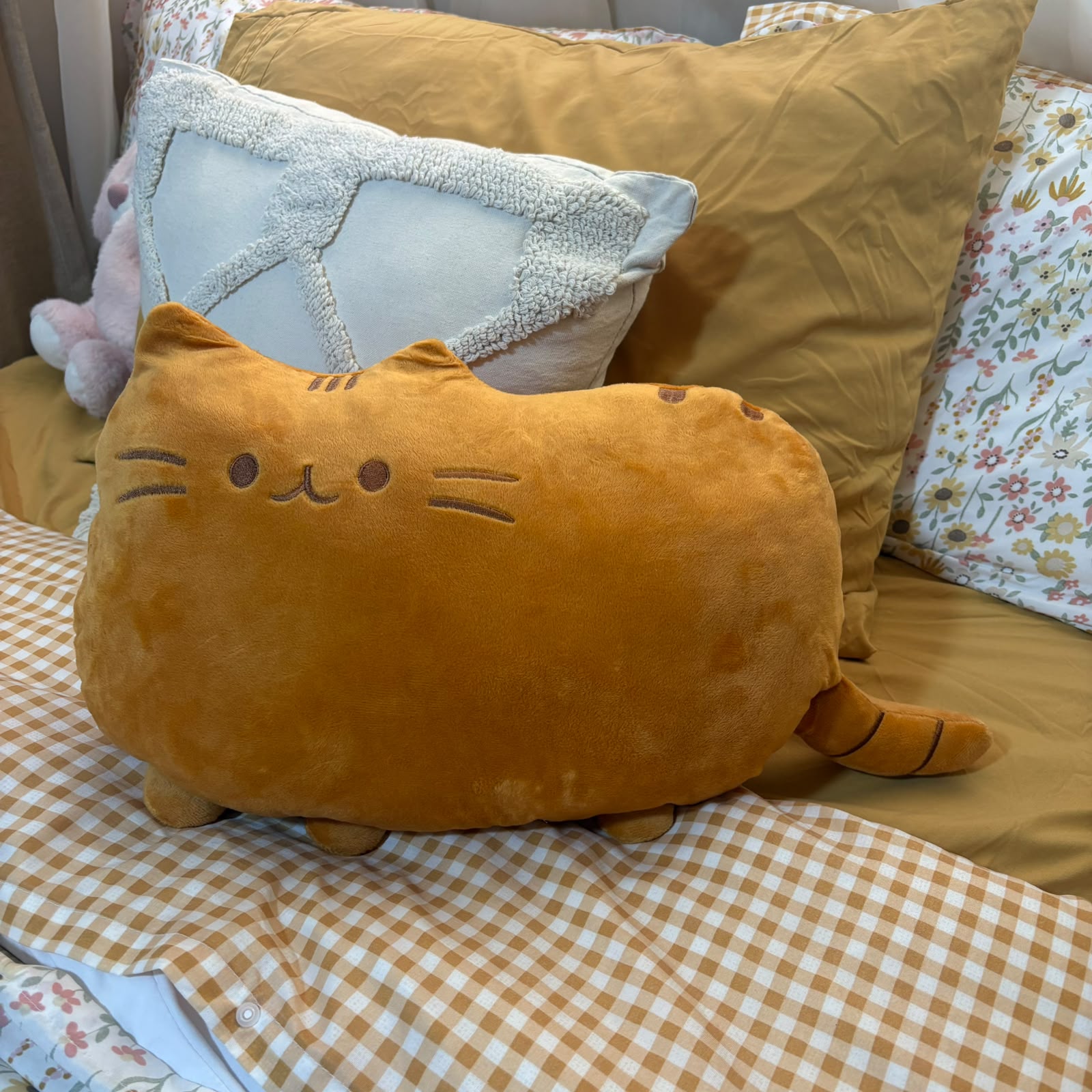 A plush cat-shaped pillow in a warm mustard-yellow color sits on a bed with a brown-and-white gingham blanket, a large tan pillow, and a light blue textured accent pillow.