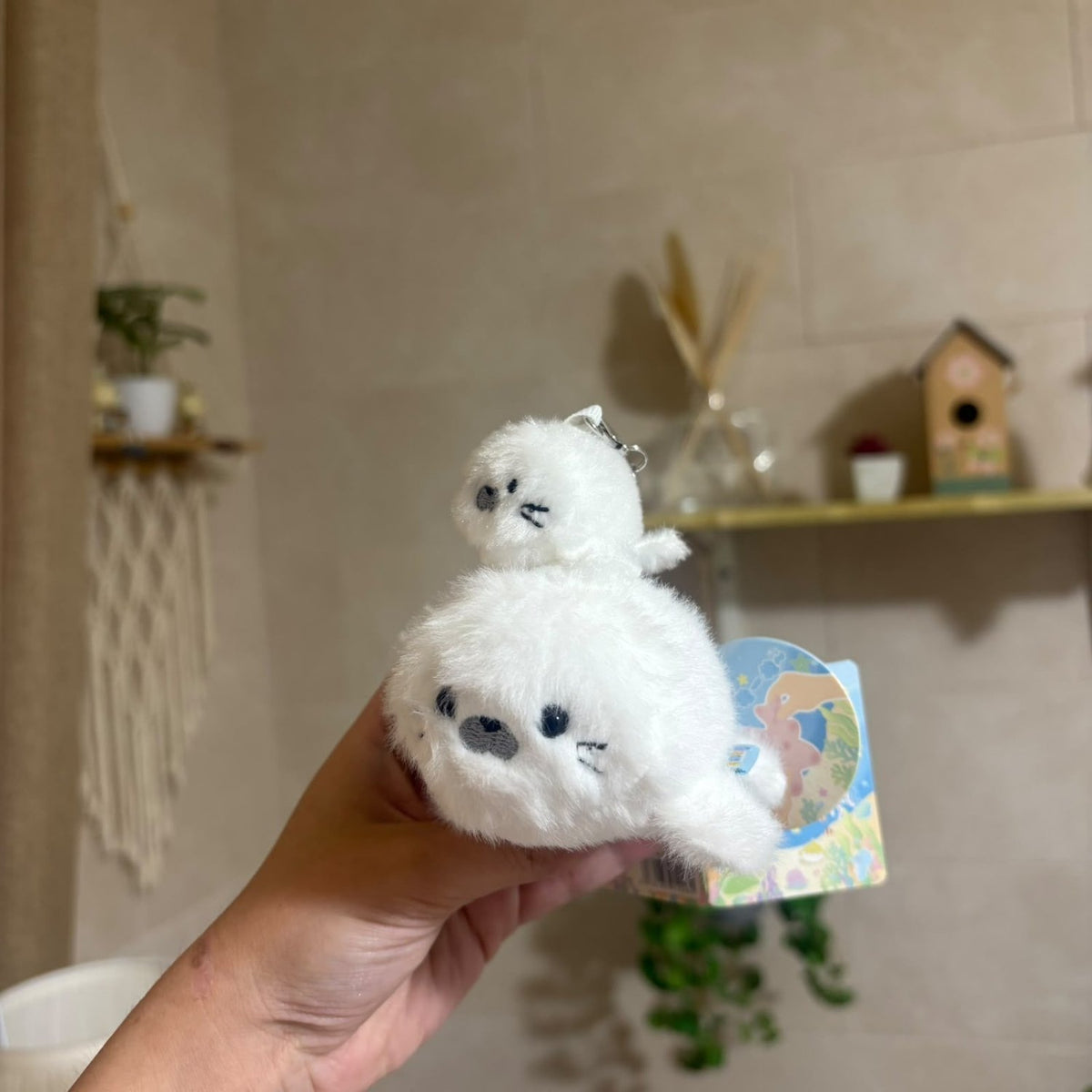 A hand holds a small white plush keychain shaped like a seal or penguin with embroidered black facial features. The plush has a small metal keyring at the top and a colorful printed backing card is attached.