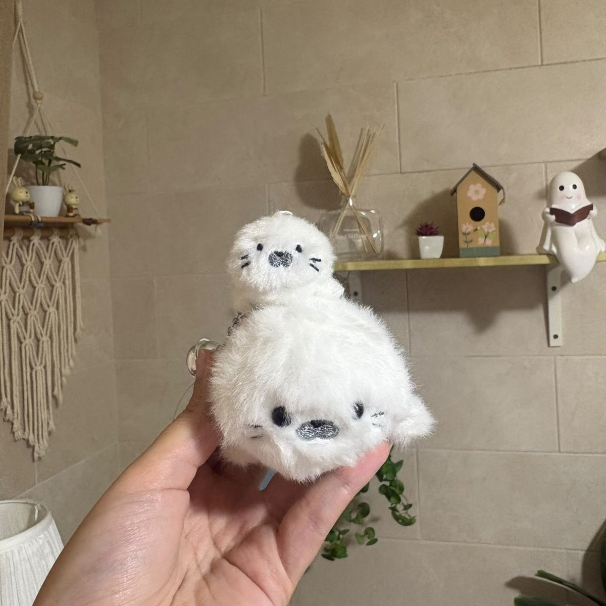 A hand holds a small white plush seal keychain in front of a beige tiled wall with shelves and decorative items in the background.