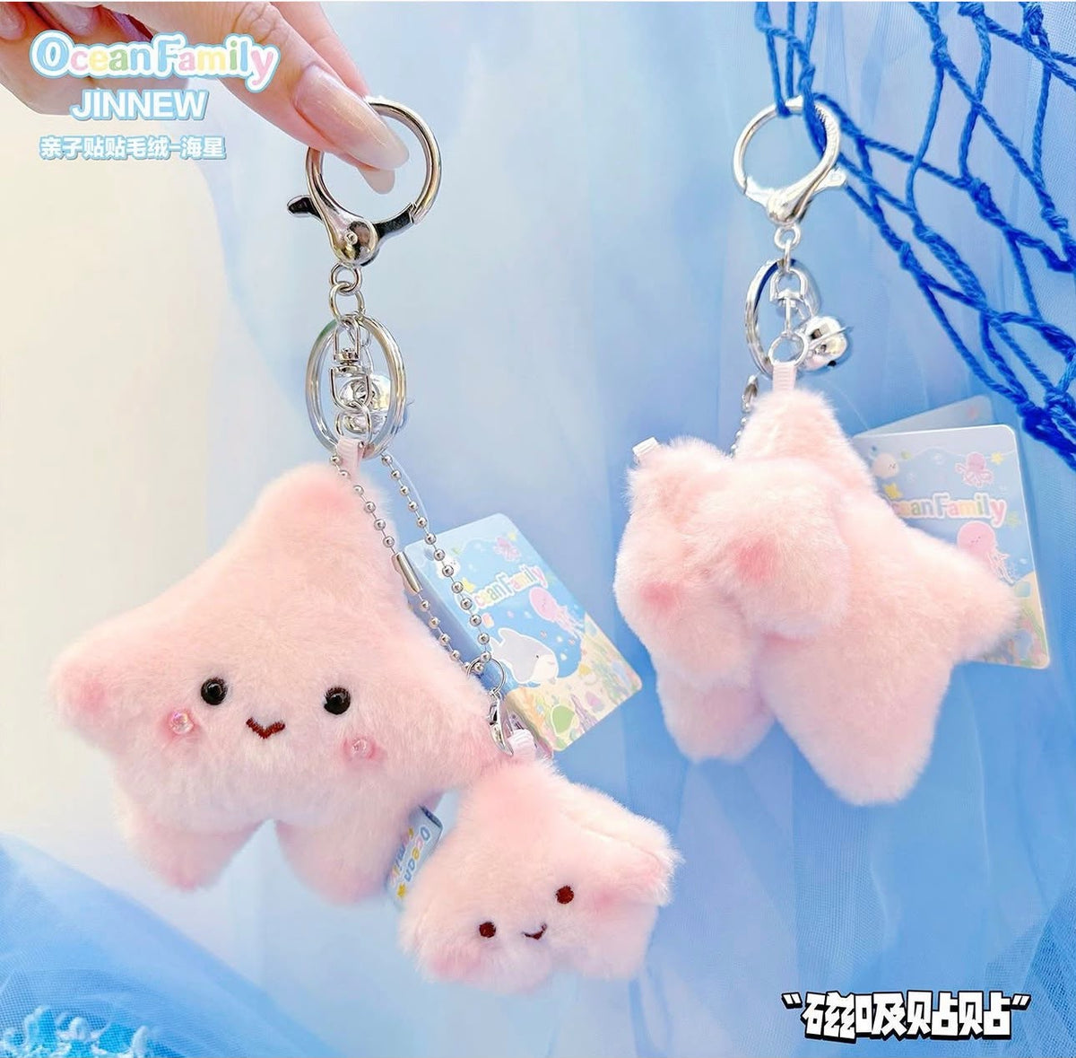 Three pink fluffy star-shaped keychains with embroidered faces are shown against a soft blue background. One keychain is held by a hand, and two others hang from metal key rings with tags attached.