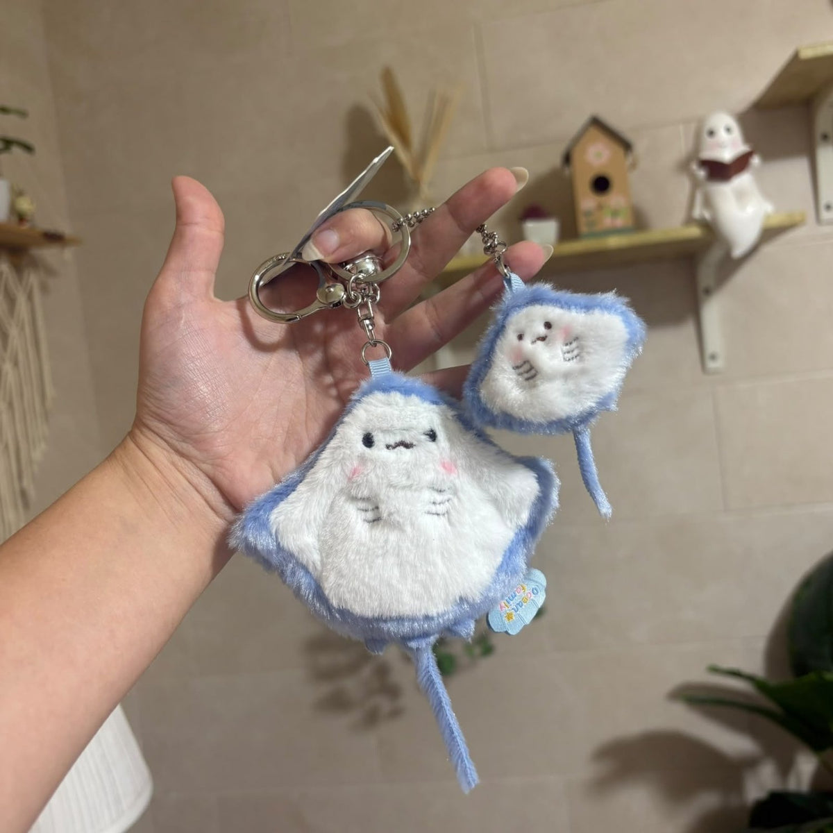 A hand holds two plush ray-shaped keychains with silver key rings in a home setting. The keychains are white with light blue fuzzy borders and embroidered facial features.