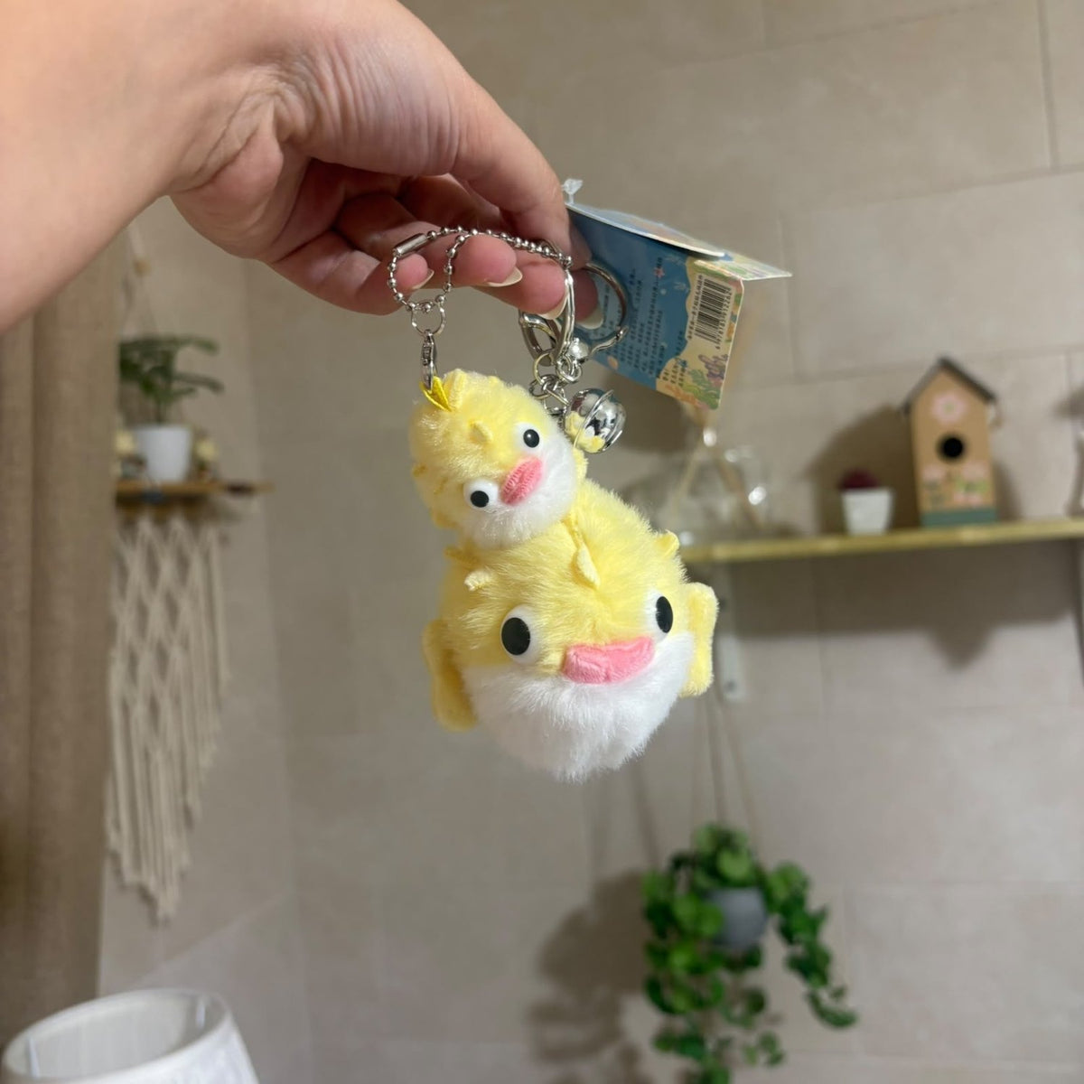 A hand holds a yellow plush duck keychain with a smaller duck on top, attached to a silver ball chain and a paper tag, against a softly blurred indoor background with shelves and plants.