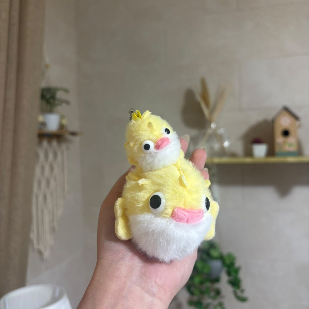 A hand holding a small yellow and white plush duck character with a pink beak and black eyes, set against a softly blurred indoor background with shelves and plants.